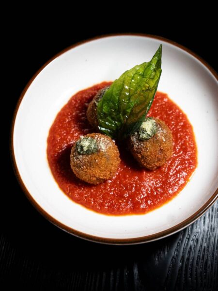 Three crispy arancini risotto balls served over rich tomato marinara sauce, garnished with pesto and a crisp basil leaf.