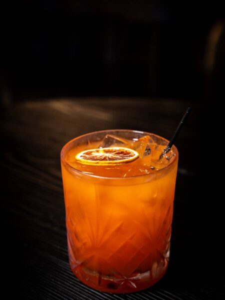 Golden Coast cocktail over ice garnished with a dehydrated orange wheel on a dark wooden bar counter.