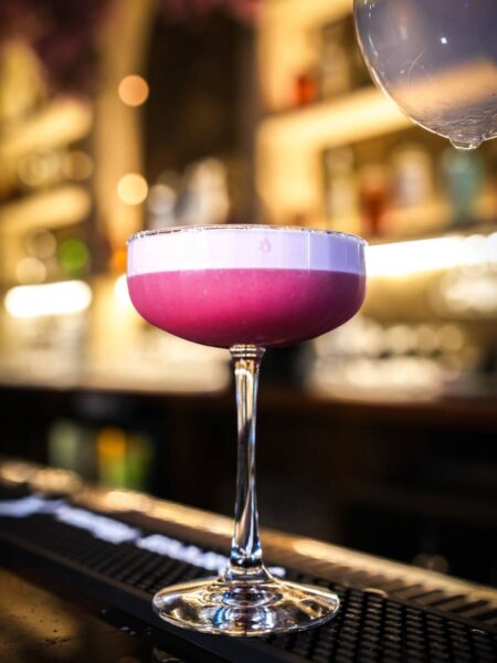 Magenta pink Hibiscus Blossom cocktail topped with silky white foam served in a coupe glass at the bar.