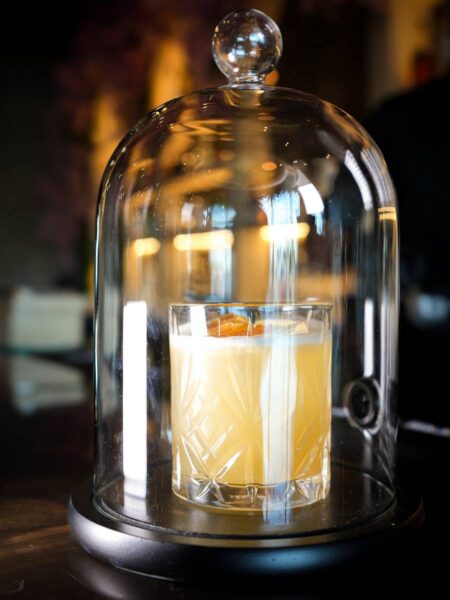 God Father cocktail encased under a glass cloche bell jar dome on a bar counter at Coca Bar & Lounge.